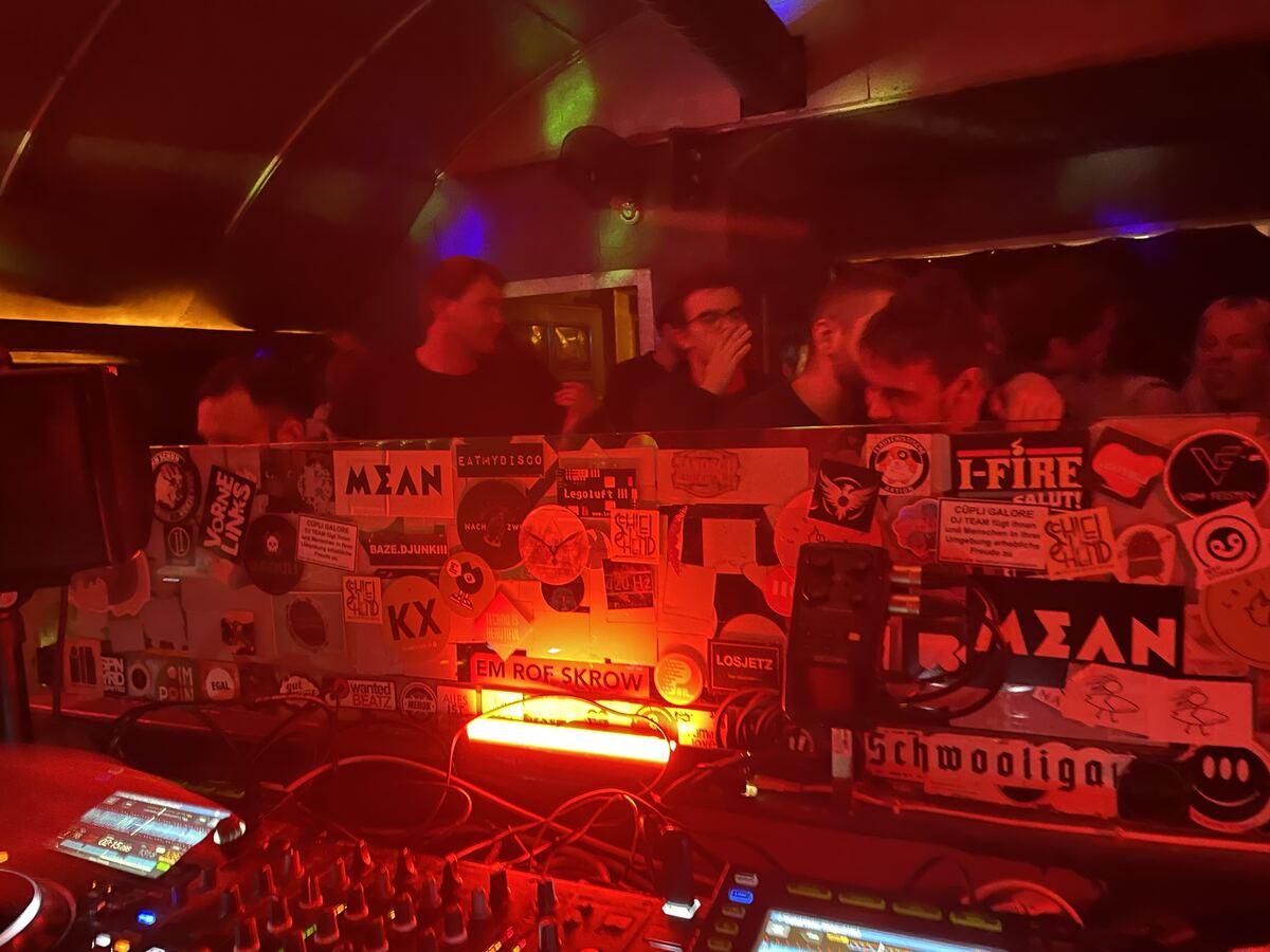 DJ booth in a club room under red light, stickers on the wall behind