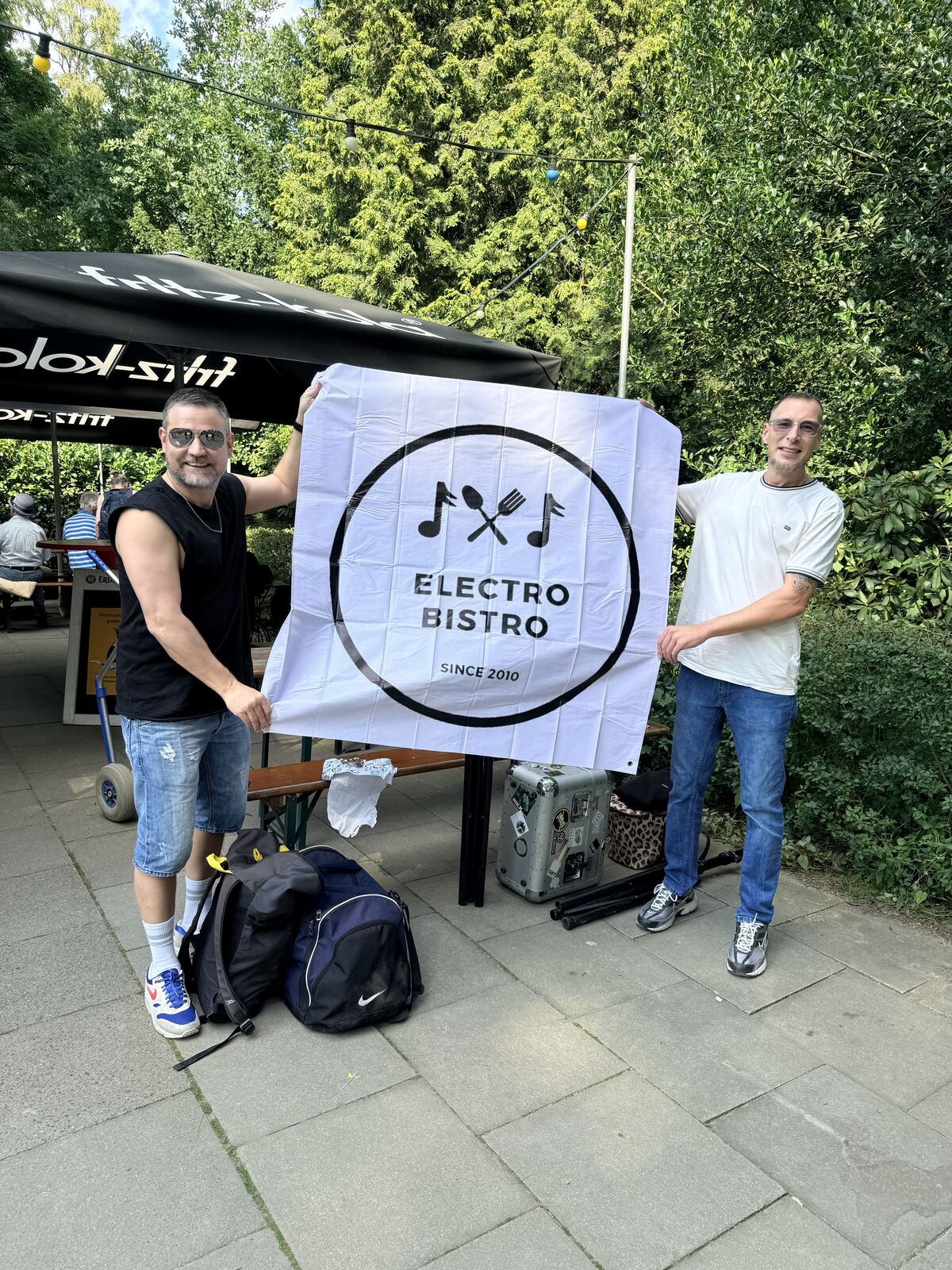 Two people holding up an Electro Bistro flag, trees and a stage behind